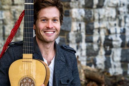 A smiling person outdoors, holding a guitar with a red strap. They are wearing a gray jacket. The background features a stone wall.