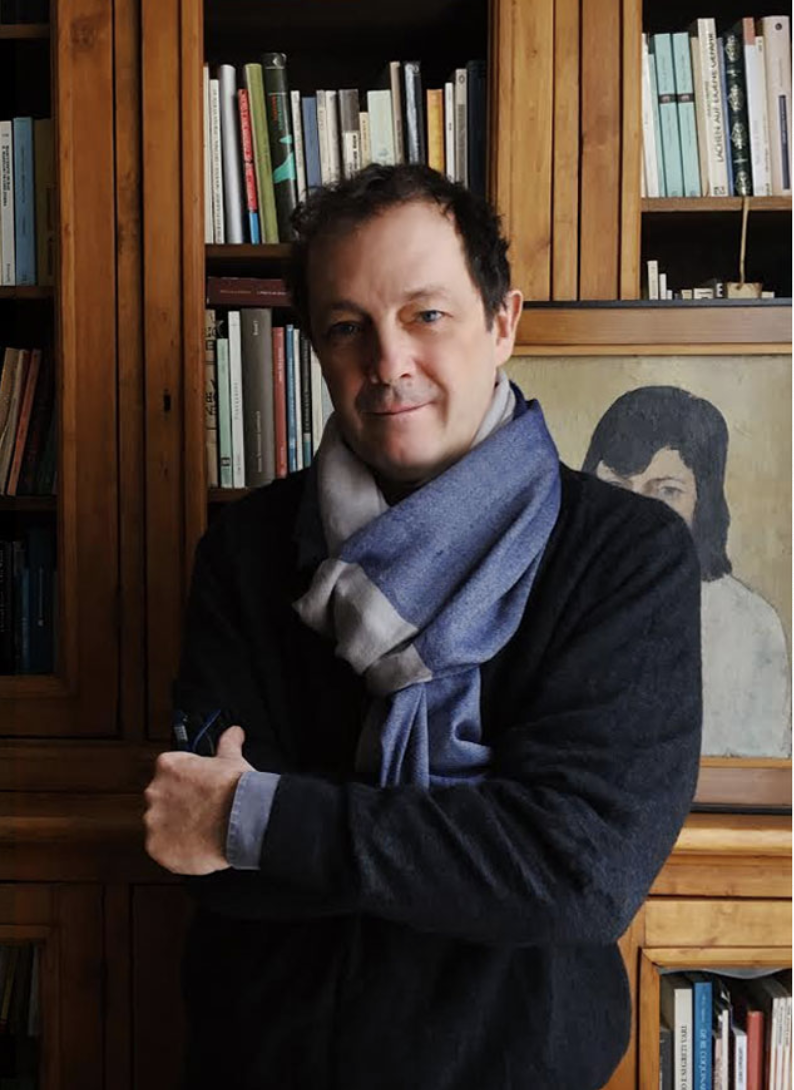 A person stands in front of a wooden bookshelf, wearing a dark sweater and a light blue scarf. They have their arms crossed and are in a cozy, book-filled room. There is a painting of another figure on the shelf behind them.