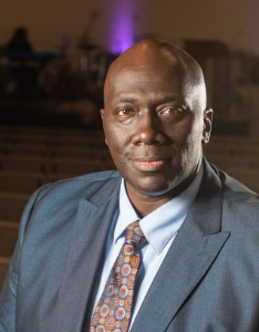 A person wearing a blue suit and patterned tie is seated indoors, facing the camera. The background is softly blurred, with subtle lighting creating a professional ambiance.