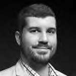 Portrait of a man with a beard, wearing a light-colored jacket and checkered shirt, smiling slightly. The background is dark, highlighting his face. The image is in black and white.