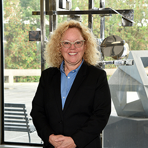 A person with curly blonde hair wearing glasses and a black suit stands indoors, smiling. Behind them, theres a window with a view of greenery and geometric art pieces.
