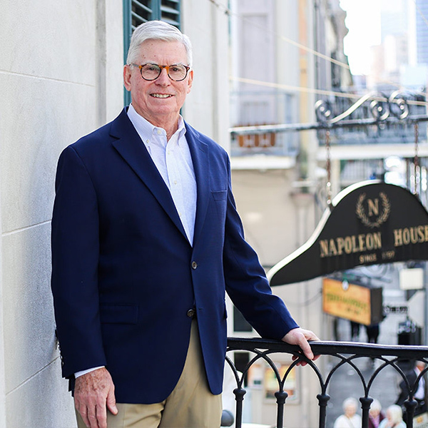 An older man wearing a navy blazer, white shirt, and khaki pants stands by a railing on a balcony. He is smiling, and a sign reading Napoleon House is visible in the background.
