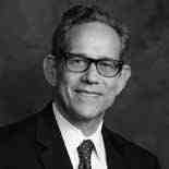 A grayscale portrait of a person wearing glasses and a suit with a tie. They have short hair and are smiling slightly. The background is a simple, dark textured backdrop.