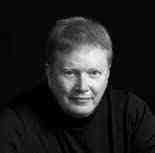 A black and white portrait of a person with short hair wearing a dark turtleneck sweater, looking directly at the camera against a dark background.