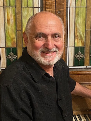 A smiling man with a white beard and short hair is wearing a black shirt. He is sitting in front of a wooden panel with stained glass details.