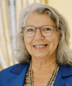 A smiling older woman with long gray hair and glasses, wearing a blue blazer and a beaded necklace, stands in front of a softly blurred background.