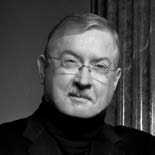 A grayscale photo of a man wearing glasses, a black jacket, and a black turtleneck sweater. He has short hair and is standing against a dark, textured background. The lighting emphasizes his facial features.