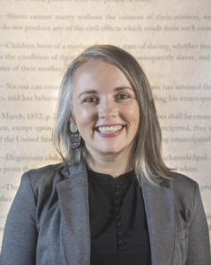 A smiling person with shoulder-length gray hair wears a blazer and earrings. They are standing in front of a wall with blurred historical text.