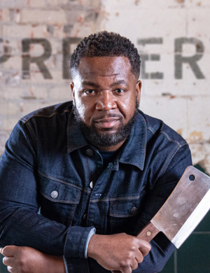 A person with short curly hair and a beard is holding a large cleaver. They are wearing a denim jacket and standing against a rustic wall with faded lettering.