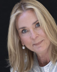 A person with long blonde hair is smiling softly, wearing a silver top and pearl earrings, set against a dark background.