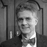 A black and white photo of a man with short curly hair wearing a bow tie and a suit jacket. He is standing in front of a wooden cabinet with decorative handles.