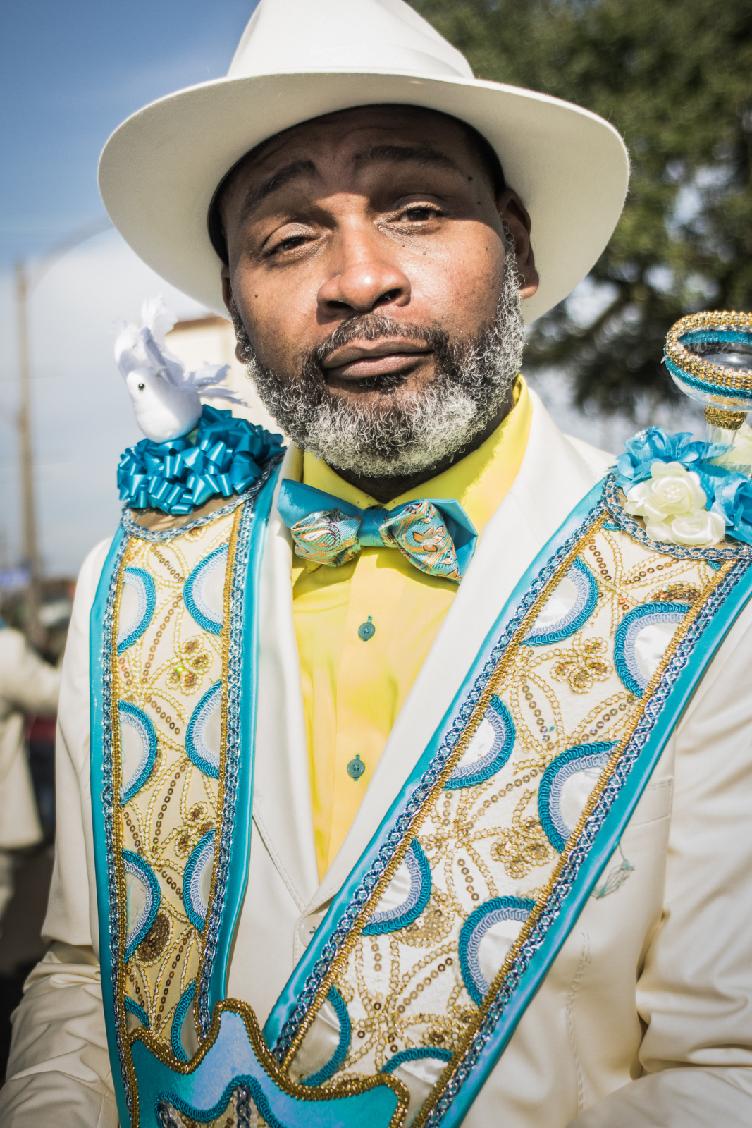 A man with a salt-and-pepper beard and a confident expression wears an elegant white hat and a matching suit adorned with intricate blue and gold embroidery. A decorative sash drapes over his shoulders, featuring a small white dove figurine and ribbon embellishments.