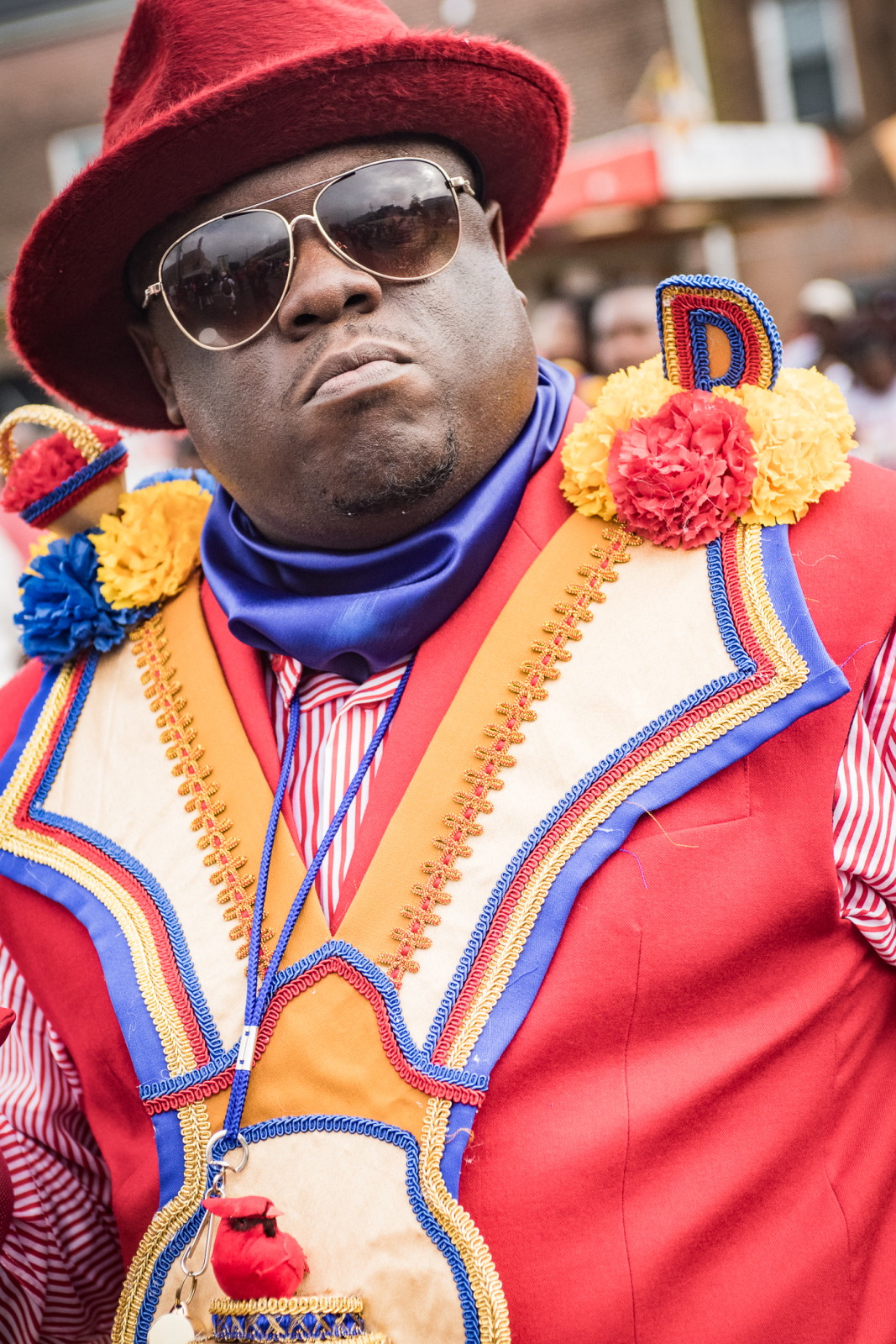 A man wearing a red hat and aviator sunglasses exudes confidence in an elaborate red, blue, and gold outfit. His embroidered sash is adorned with vibrant fabric flowers and intricate beadwork, reflecting the bold style of a New Orleans second-line parade.