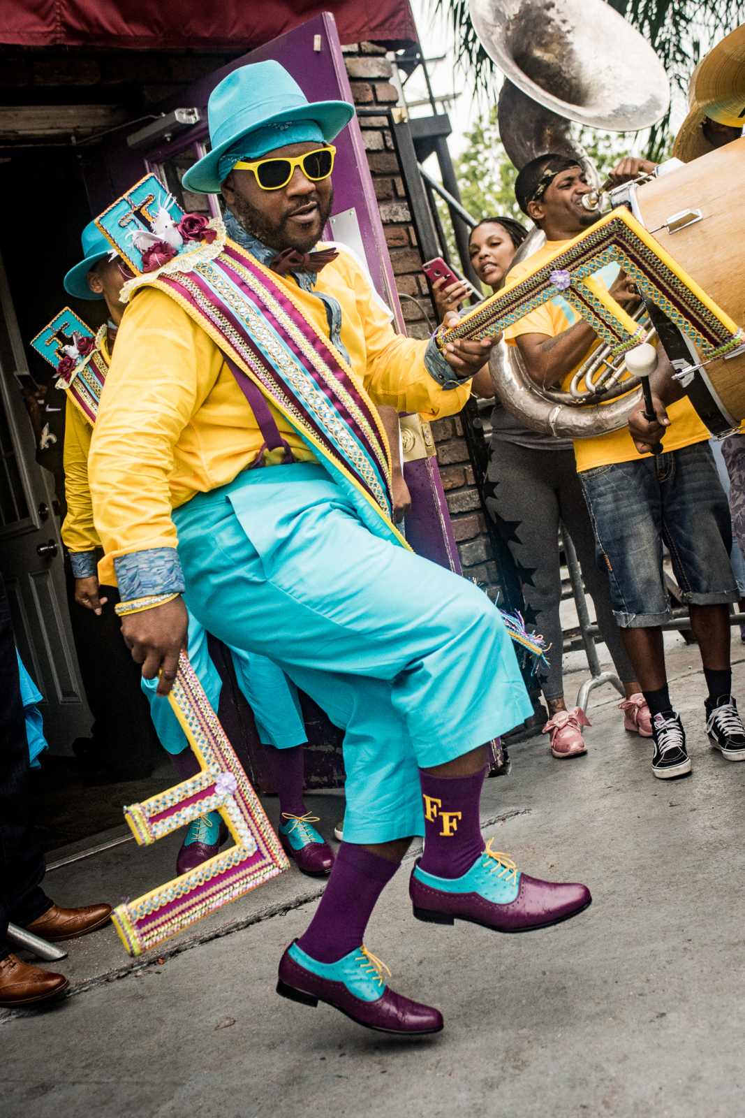 Derrick Santa Cruz dances energetically in a bright yellow and turquoise outfit, holding a decorated letter "F" prop, as a brass band plays behind him during a lively street parade.