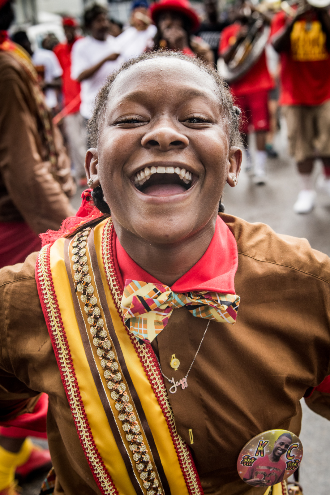 Close-up of Jazz Henry laughing during a parade, wearing a colorful bowtie, a decorative sash, and a button with "KC" on it, as people in bright outfits and brass instruments fill the street behind them.