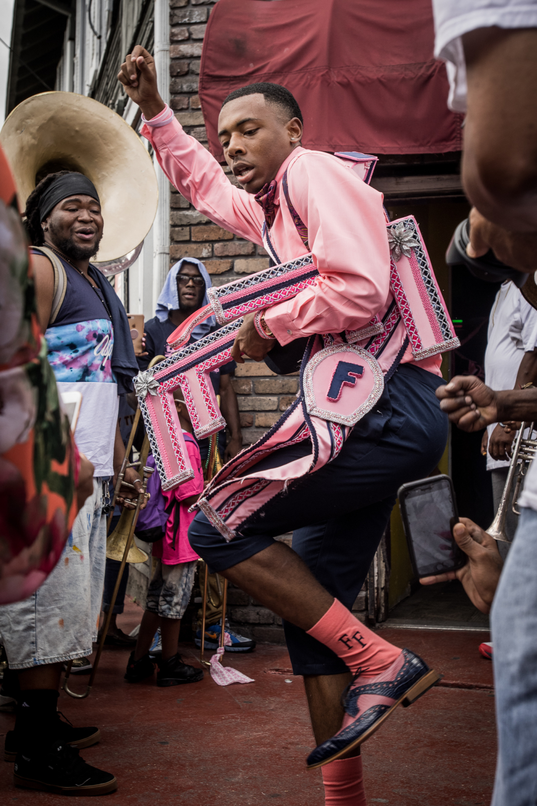 Jeremy Horn, wearing a pink shirt and navy pants and dancing, wears intricate pink and silver sashes, as brass musicians and a crowd surround him in celebration.
