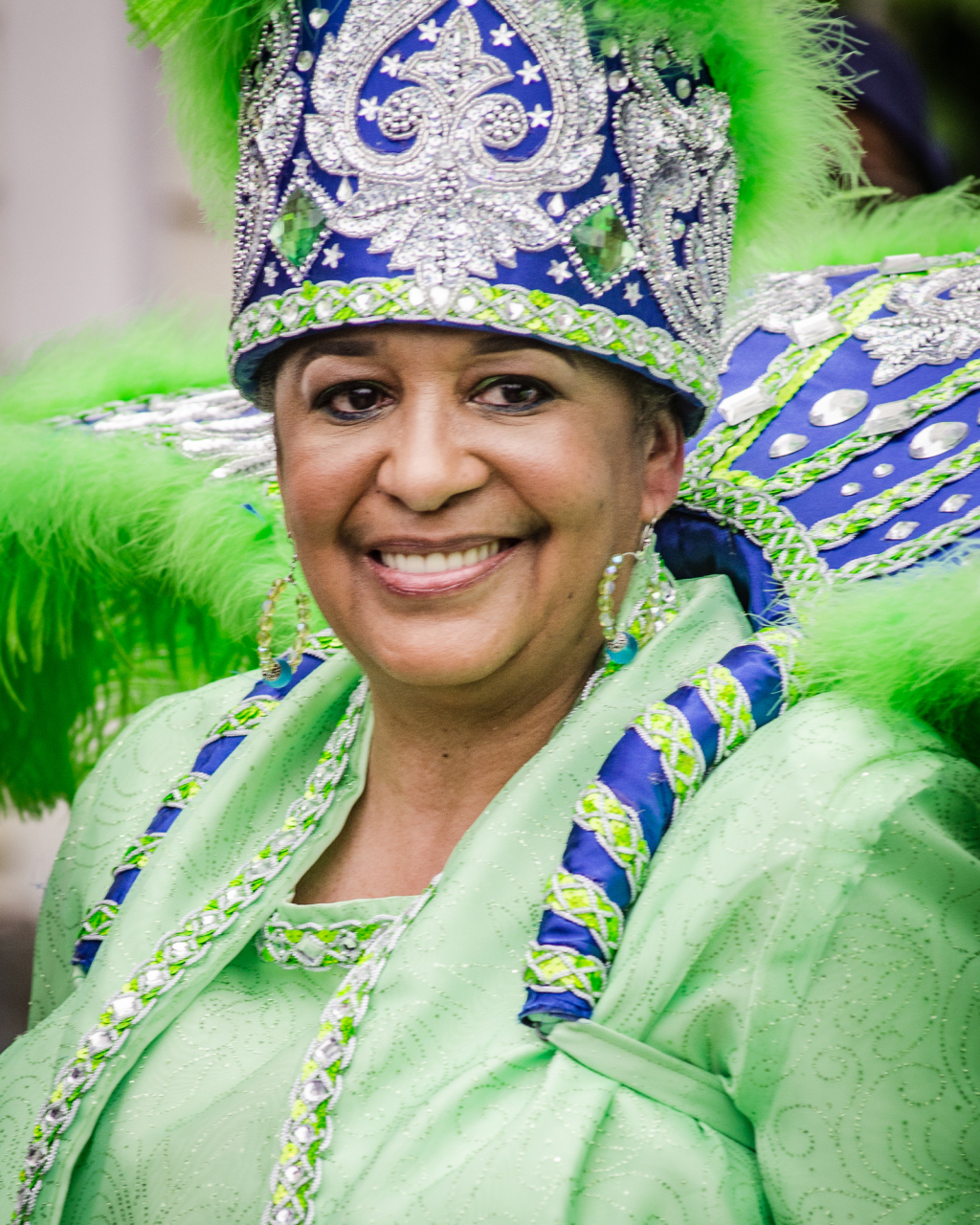 A person smiling while wearing a colorful, ornate costume with a large headdress adorned with green feathers, blue and silver patterns. The outfit is bright green with matching details. They appear to be participating in a festive event.