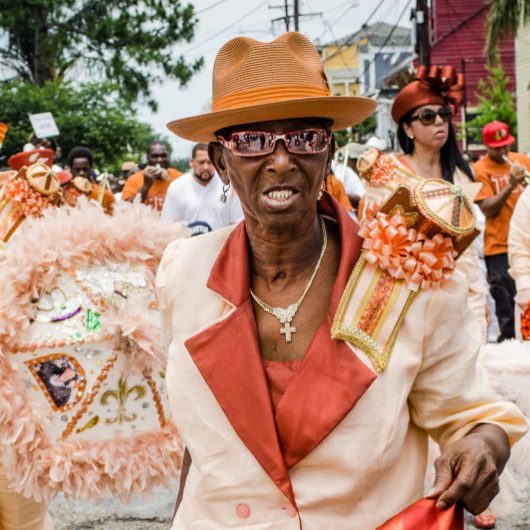 A person in stylish attire, including a peach suit and hat, participates in a parade. The crowd around them features vibrant decorations and costumes. A festive atmosphere is evident, with people enjoying the lively event.