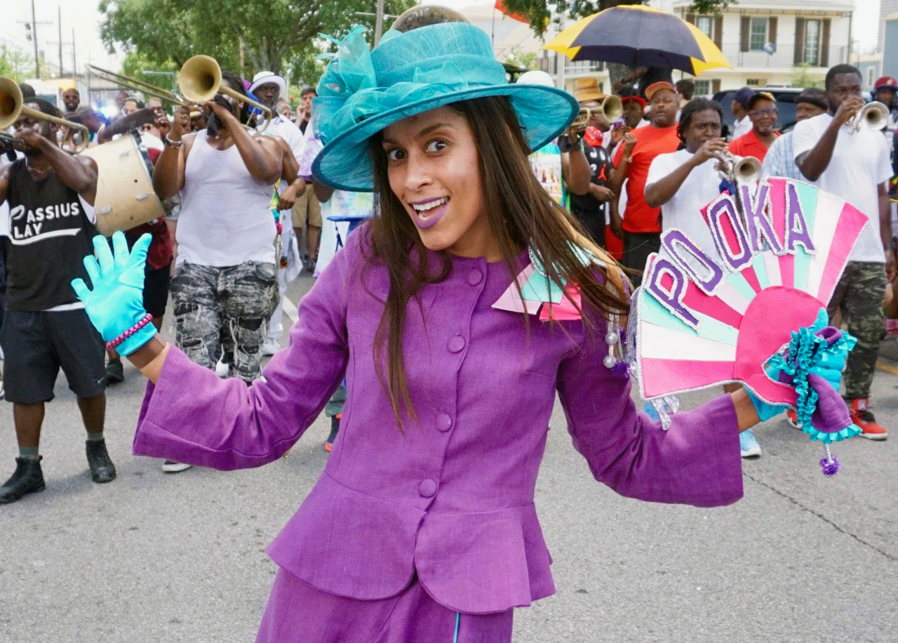 A smiling woman in a bright purple suit and teal hat dances in a street parade. She holds a colorful fan with POOKA written on it. Around her, a band plays instruments, while other people watch, some holding umbrellas.