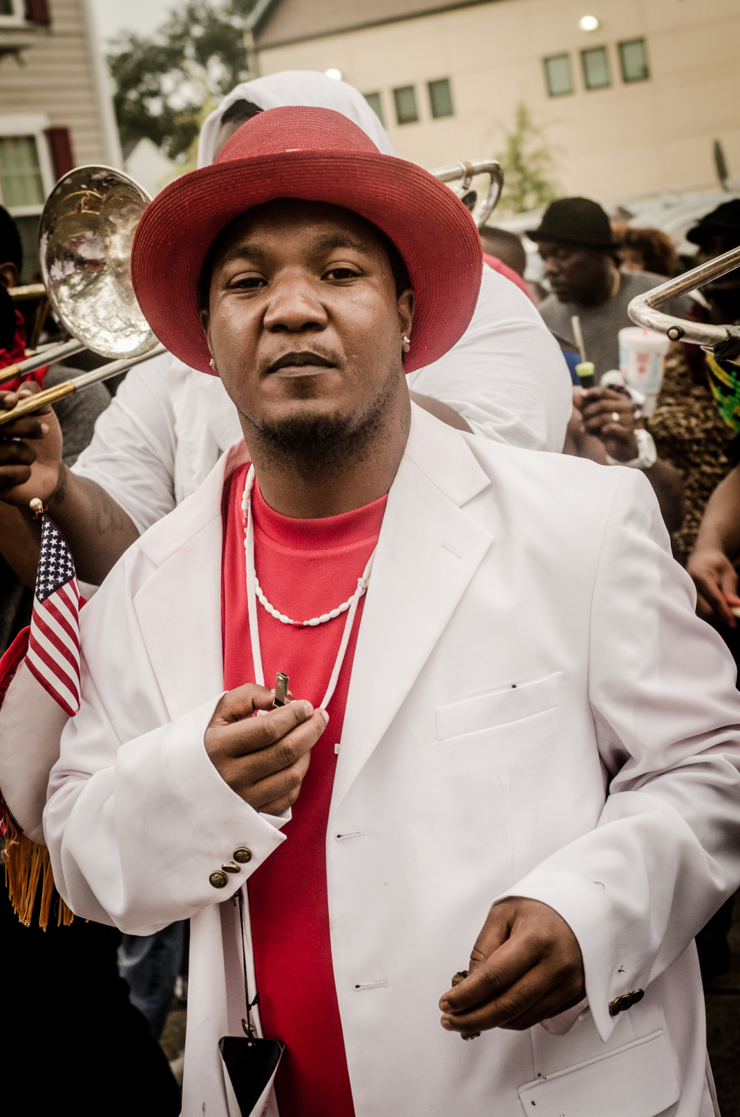 A man in a red hat, red shirt, and white suit stands confidently at a second-line parade, holding a whistle and looking directly at the camera while brass band musicians play behind him.