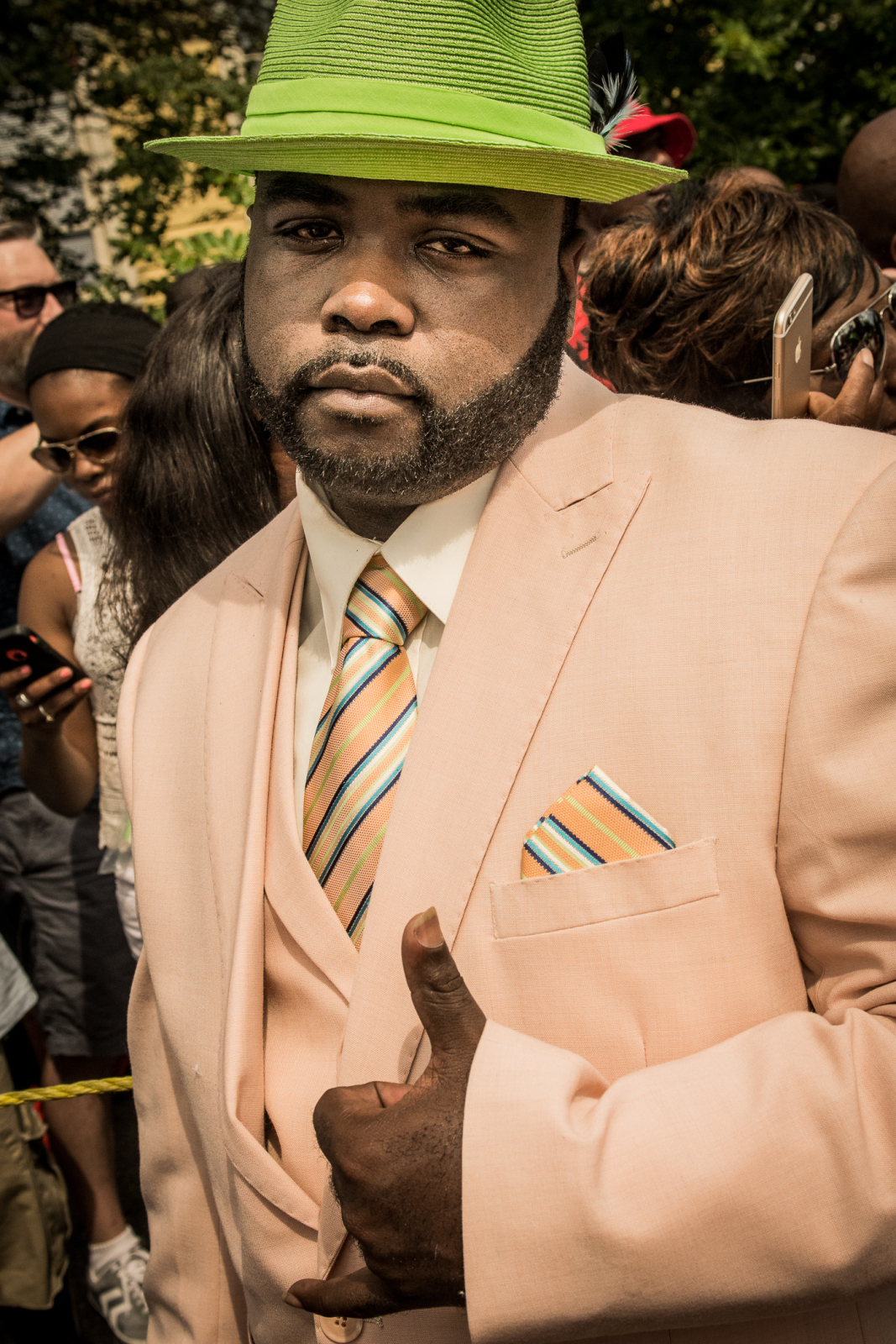 A man in a pastel peach suit and vibrant green hat gives a subtle thumbs-up at a second line parade, surrounded by a crowd.