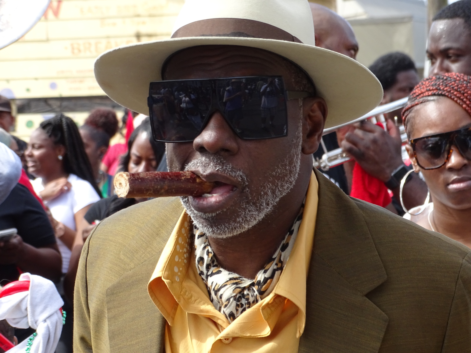 A photo shows Greg Stafford with a gray beard wearing a wide-brimmed hat, large sunglasses, and a mustard-yellow shirt with a patterned scarf. He holds a thick cigar in his mouth, surrounded by people and musicians at a parade.