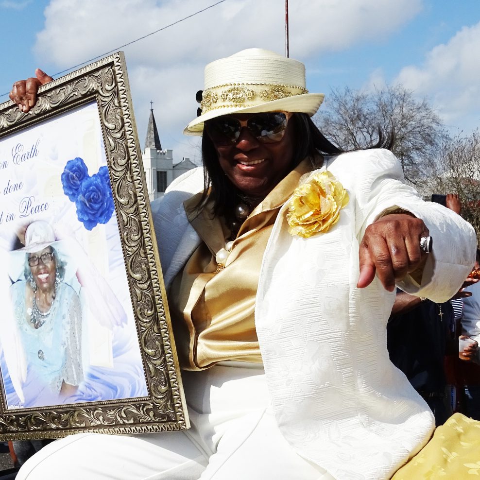 Shelia Gibson honors her mother, Naomi Gibson, at the Lady Jetsetters 30th anniversary parade. She is wearing a white suit with gold shirt and hat, and is riding in an open-top convertible.