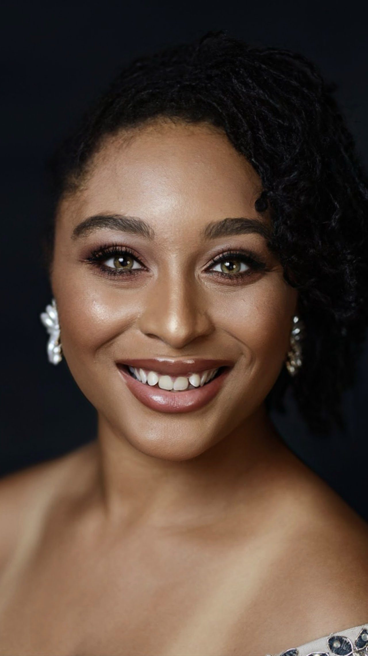 A person with shoulder-length dark hair styled to one side, wearing pearl earrings and a light-colored top, smiles warmly against a dark background.
