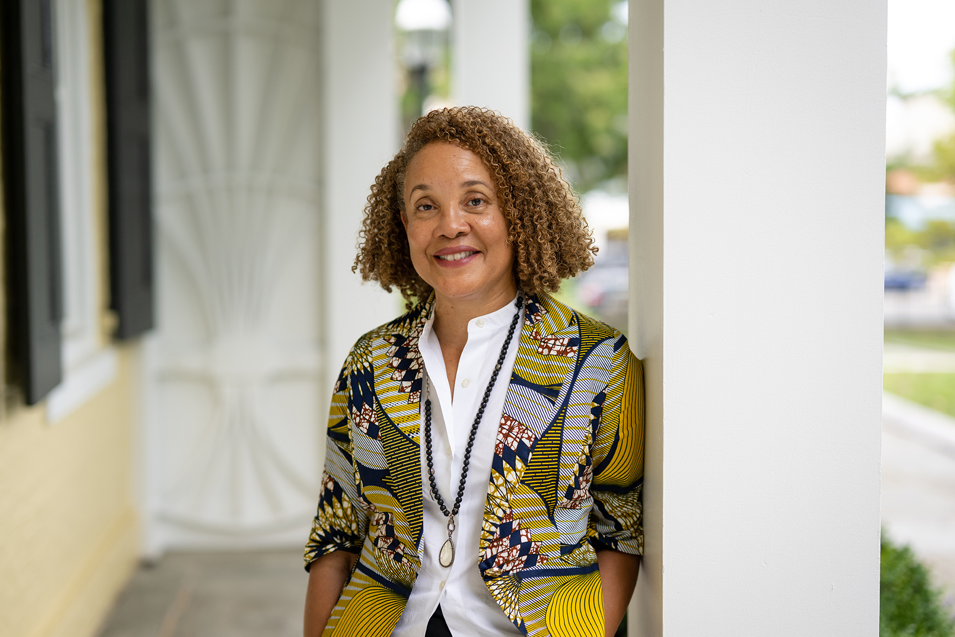 A person with curly hair stands outside, leaning against a white column. They are smiling and wearing a colorful, patterned jacket over a white shirt. The background includes a building facade and greenery.