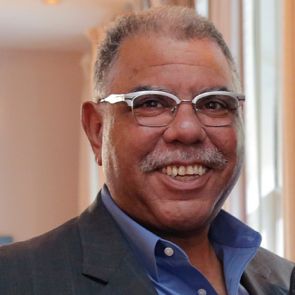 A smiling man with gray hair and a mustache is wearing glasses, a blue shirt, and a gray blazer. He stands indoors in front of a softly lit background with windows.