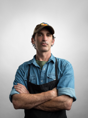 A person wearing a denim shirt, black apron, and a cap stands with arms crossed against a neutral background. They have a relaxed, confident expression.