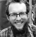 A bearded man with glasses is smiling broadly. He has short, wavy hair and is wearing a plaid shirt. The background is blurred, suggesting an outdoor setting. The image is in black and white, highlighting the mans cheerful expression.