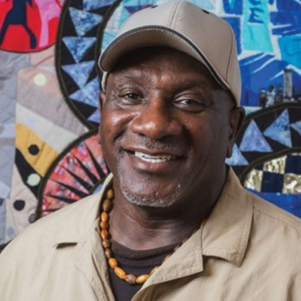 Smiling man wearing a beige cap and shirt, with a beaded necklace. Colorful abstract art is in the background.