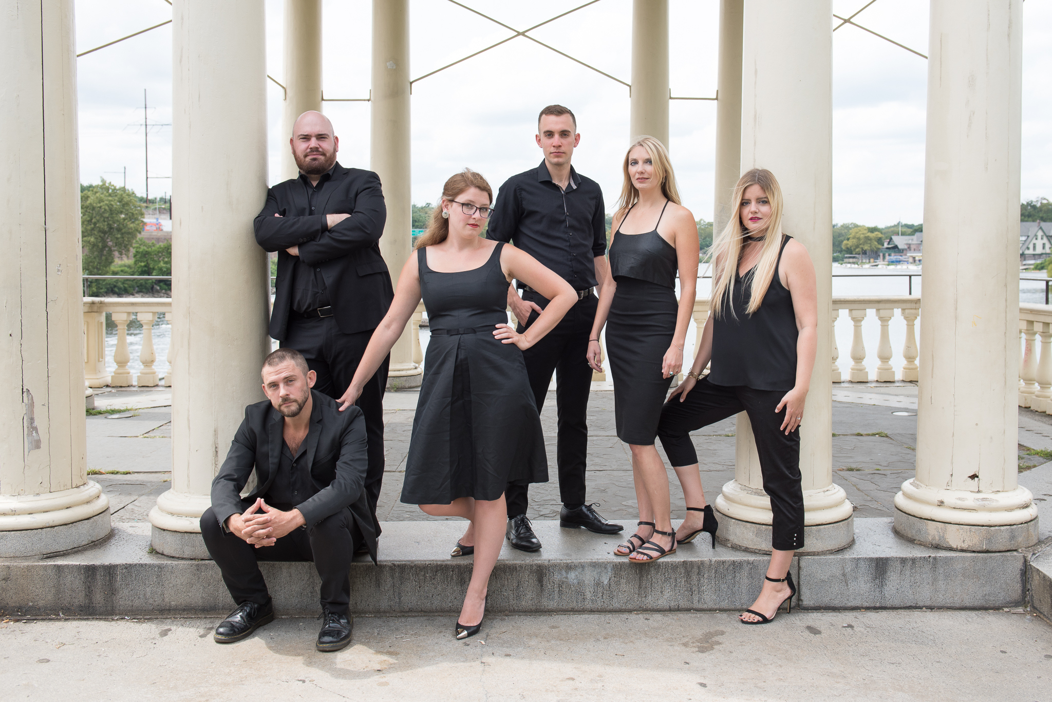 A group of six people, all dressed in black attire, pose confidently among large white columns outdoors. The background shows a stone railing and a view of trees and water.