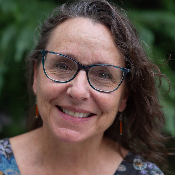 A person with glasses and long wavy hair smiles warmly at the camera. They are wearing a patterned top and orange earrings. The background is blurred greenery.