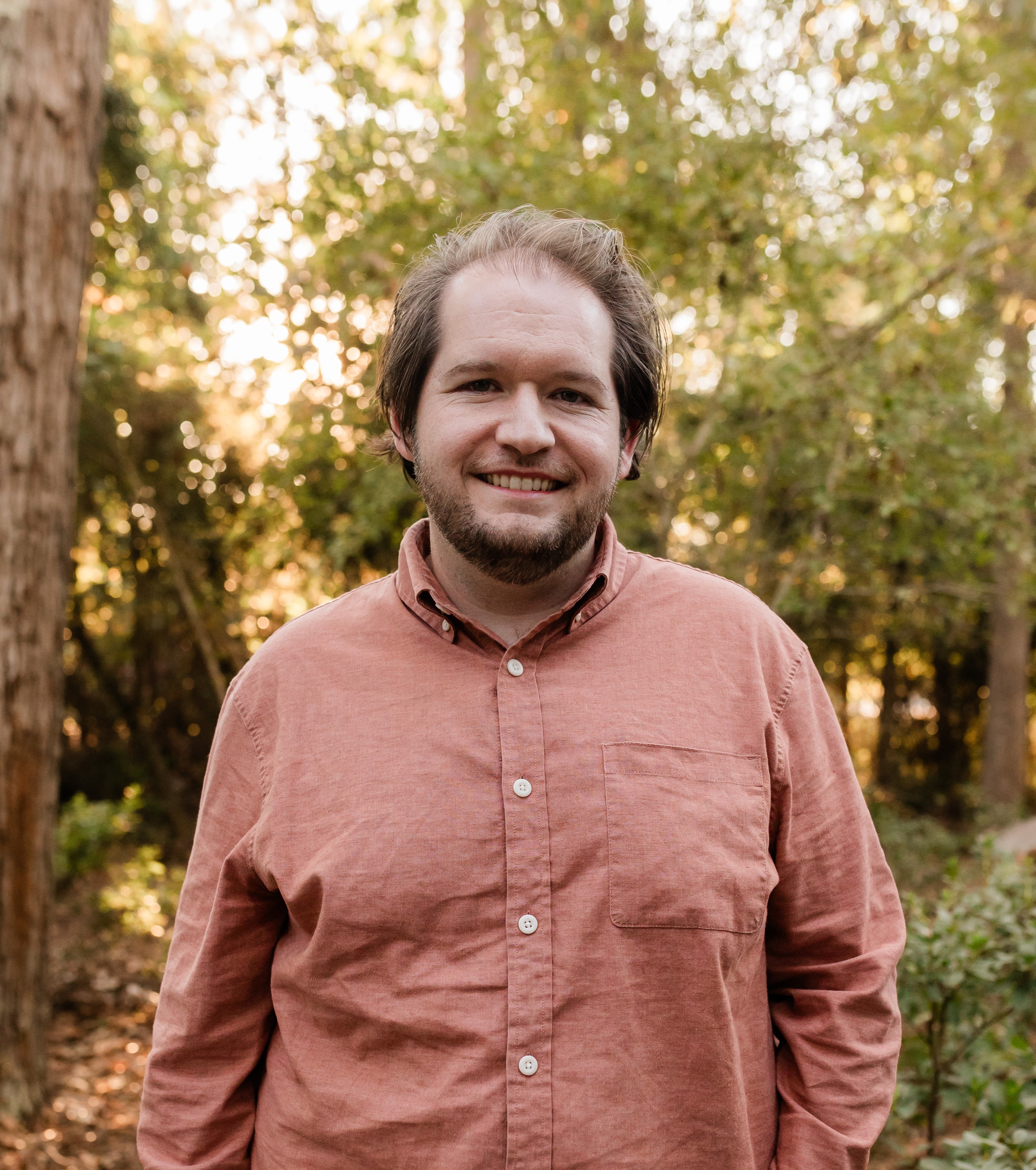 Photo of William Jones with a beard and medium-length hair standing in a forested area, wearing a salmon-colored button-up shirt. The background includes tall trees and sunlight filtering through the leaves, creating a warm and natural setting.