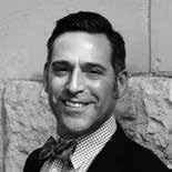 A black and white portrait of a smiling man wearing a checkered shirt, bow tie, and suit jacket. The background features a stone wall.