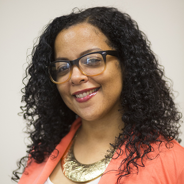 A woman with curly hair and glasses smiles at the camera, wearing a coral blazer, a white top, and a large gold necklace. The background is plain and light-colored.