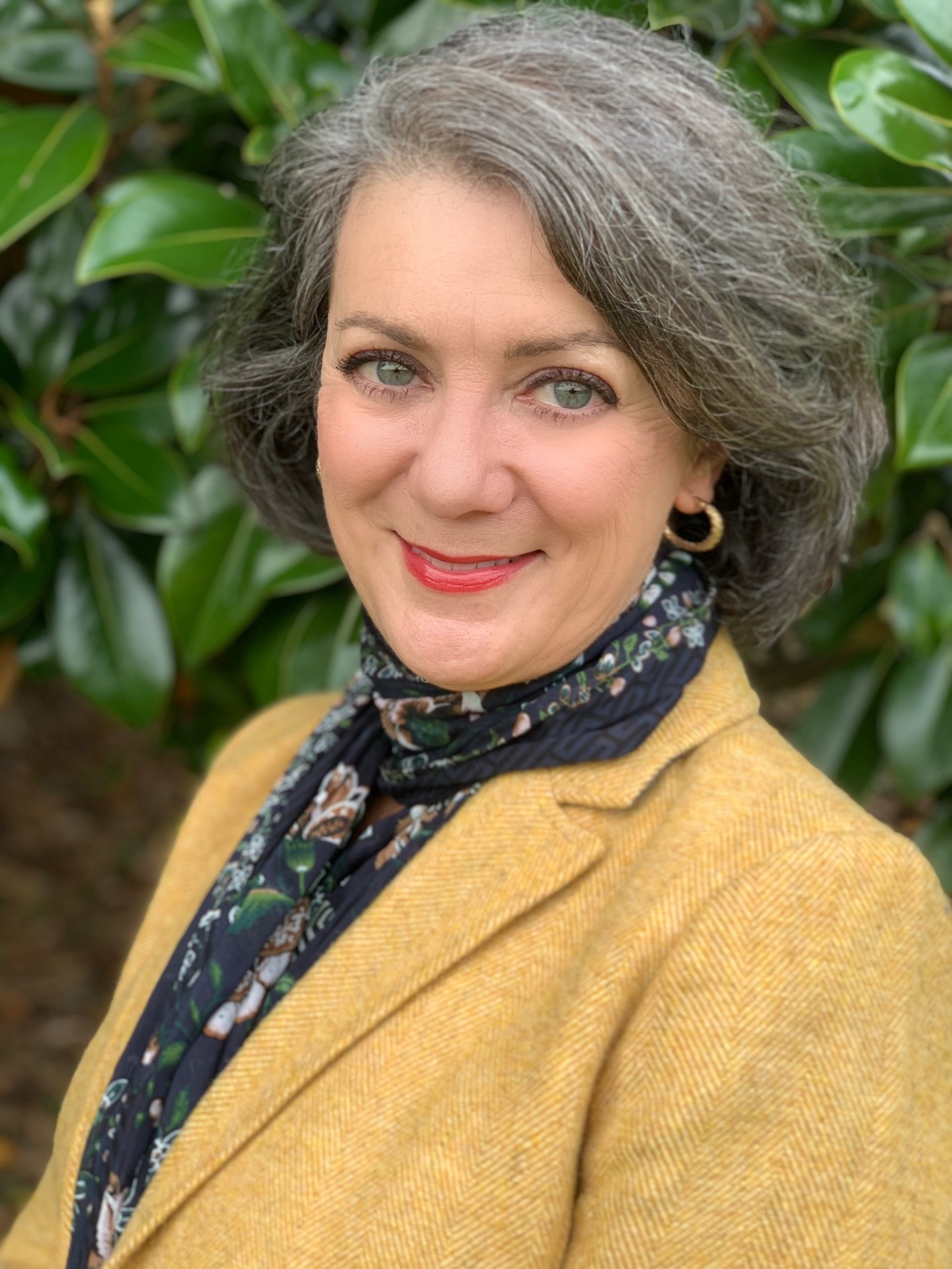 A woman with gray hair and green eyes is smiling, wearing a yellow blazer and a floral scarf. She stands in front of large green leaves outdoors.