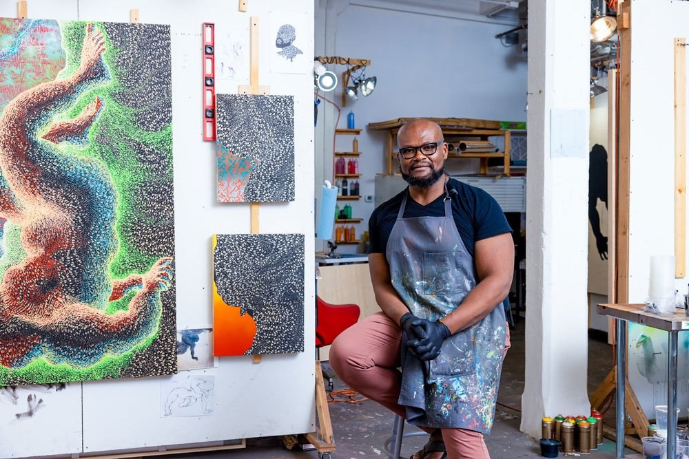 An artist wearing an apron and gloves sits on a stool in a studio, surrounded by vibrant, abstract paintings. The paintings feature colorful, dynamic patterns. The studio is bright, with various art materials visible in the background.