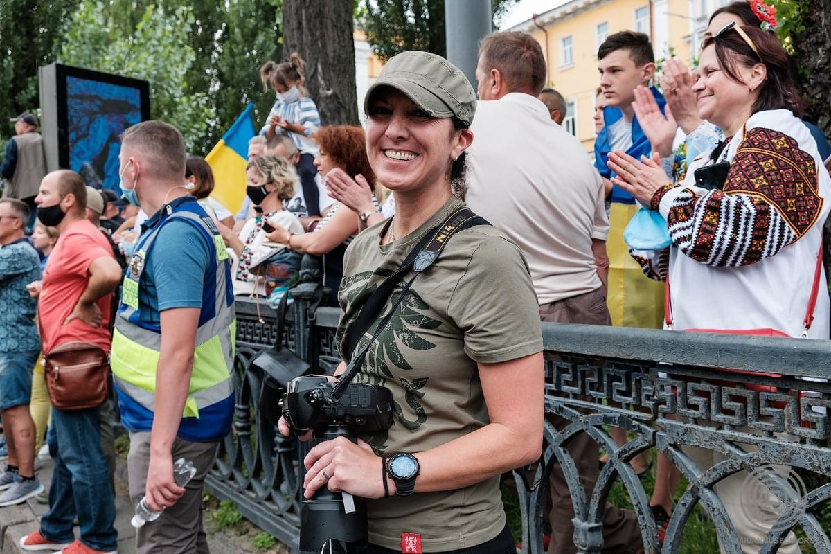 A smiling woman in a green hat and shirt, holding a camera, stands in front of a crowd. Behind her, people are clapping and holding Ukrainian flags. The scene is outdoors with trees and a building in the background.