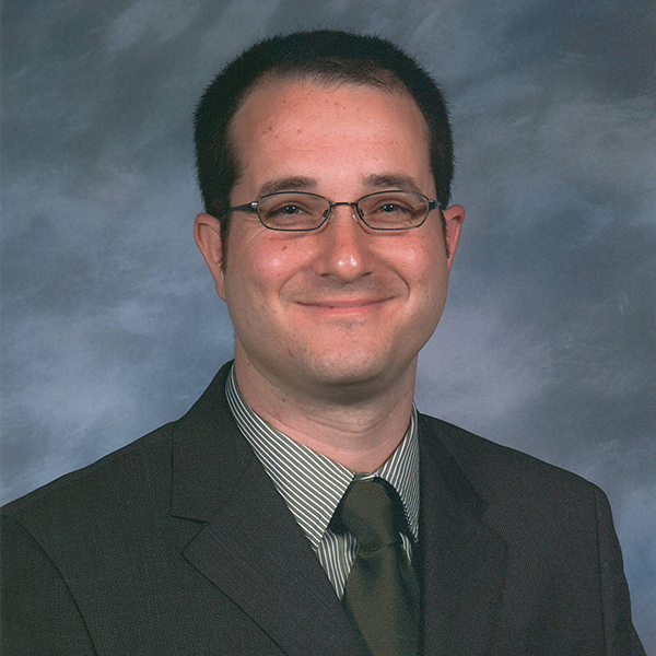 A man in a suit and tie is smiling in front of a gray and blue mottled background. He is wearing glasses and has short dark hair.