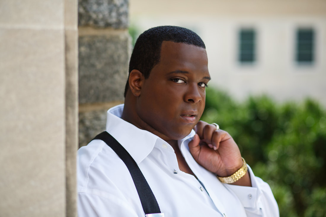A person stands outdoors, leaning against a stone wall. They are wearing a white shirt with black suspenders and a gold watch, with greenery and buildings blurred in the background. The person has a thoughtful expression.