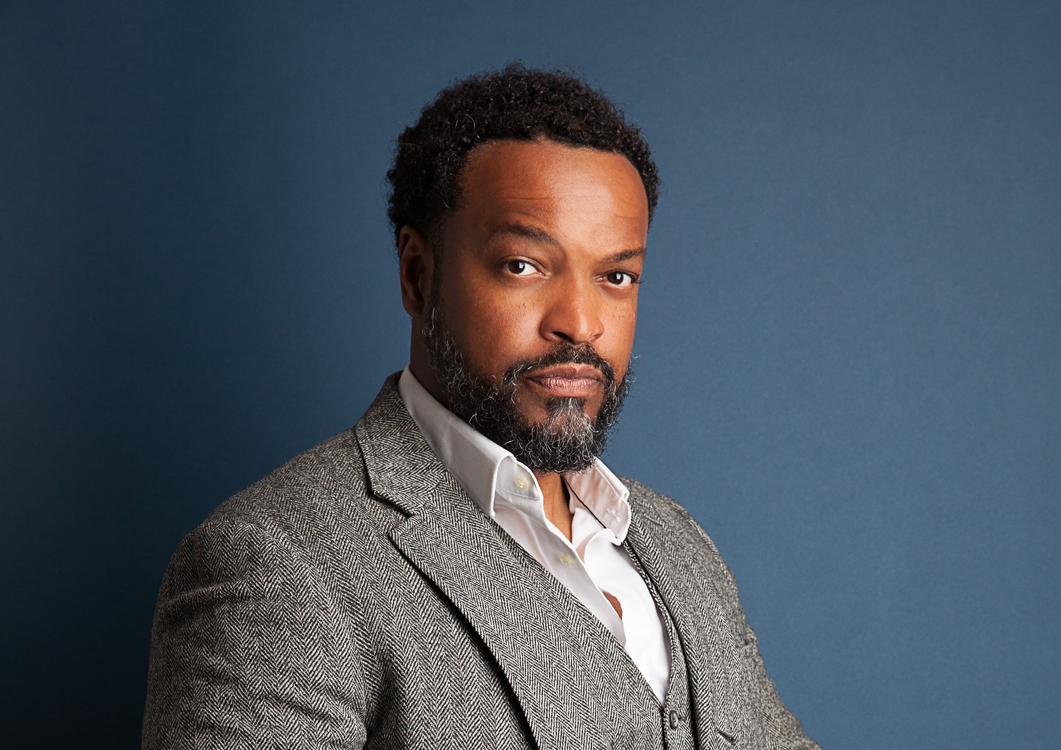 A man with short curly hair and a beard is wearing a gray tweed suit over a white collared shirt. He is posing against a dark blue background, looking confidently at the camera.