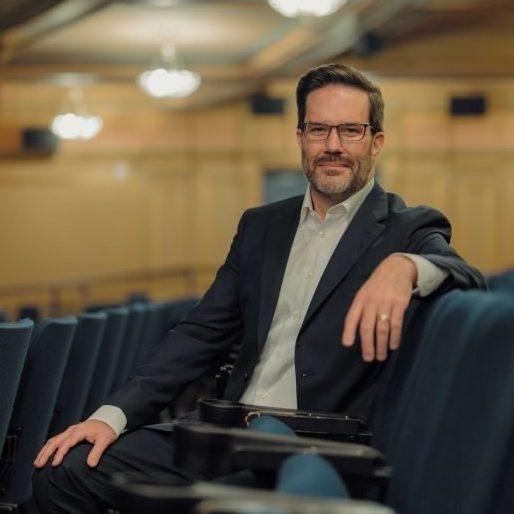 A man in a suit sits comfortably in a theater filled with rows of empty blue seats. The background features ornate lighting fixtures and elegant architectural details, indicating a classic theater setting.