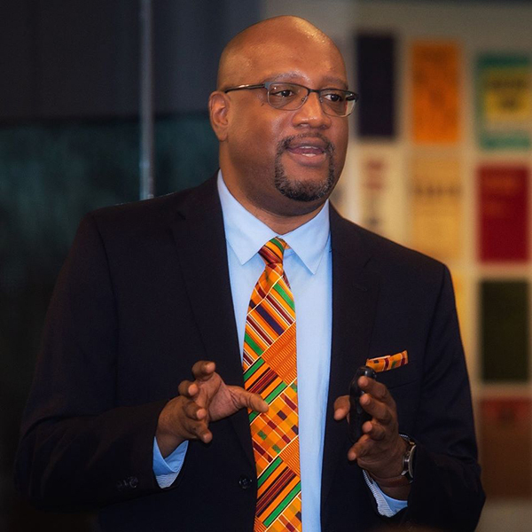 A person wearing glasses and a suit with a colorful patterned tie speaks and gestures with hands. There are colorful book covers on the wall in the background.