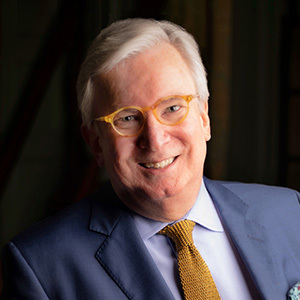 A man with gray hair and glasses smiles while wearing a blue suit, white shirt, and a yellow tie. The background is dark and blurred.