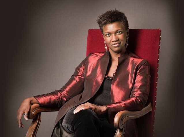 A person sits in a red velvet armchair against a gray background. They are wearing a shiny, dark red blazer over a black top, with their legs crossed and hands resting on the chairs arms. They have a short hairstyle and are looking confidently at the camera.
