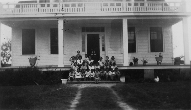 A group of children and a few adults pose on the steps of a large two-story house with a porch. The house has several windows and potted plants. A chicken is visible on the grass in the front yard. The scene is in black and white.