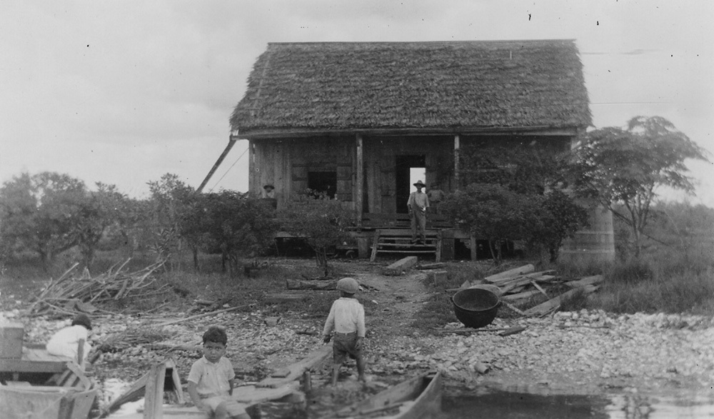 A rustic wooden house with a thatched roof stands by a lakeside. Two children are playing near a canoe in the water, while two adults are on the porch. The surrounding area is lush with trees and bushes, and the sky is overcast.